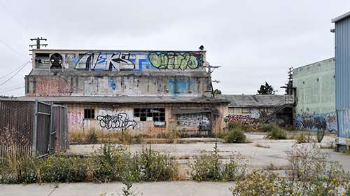750 Gilman Street before redevelopment: a low graffiti-covered building behind chain-link fencing, weeds pushing through cracked asphalt under an overcast sky.