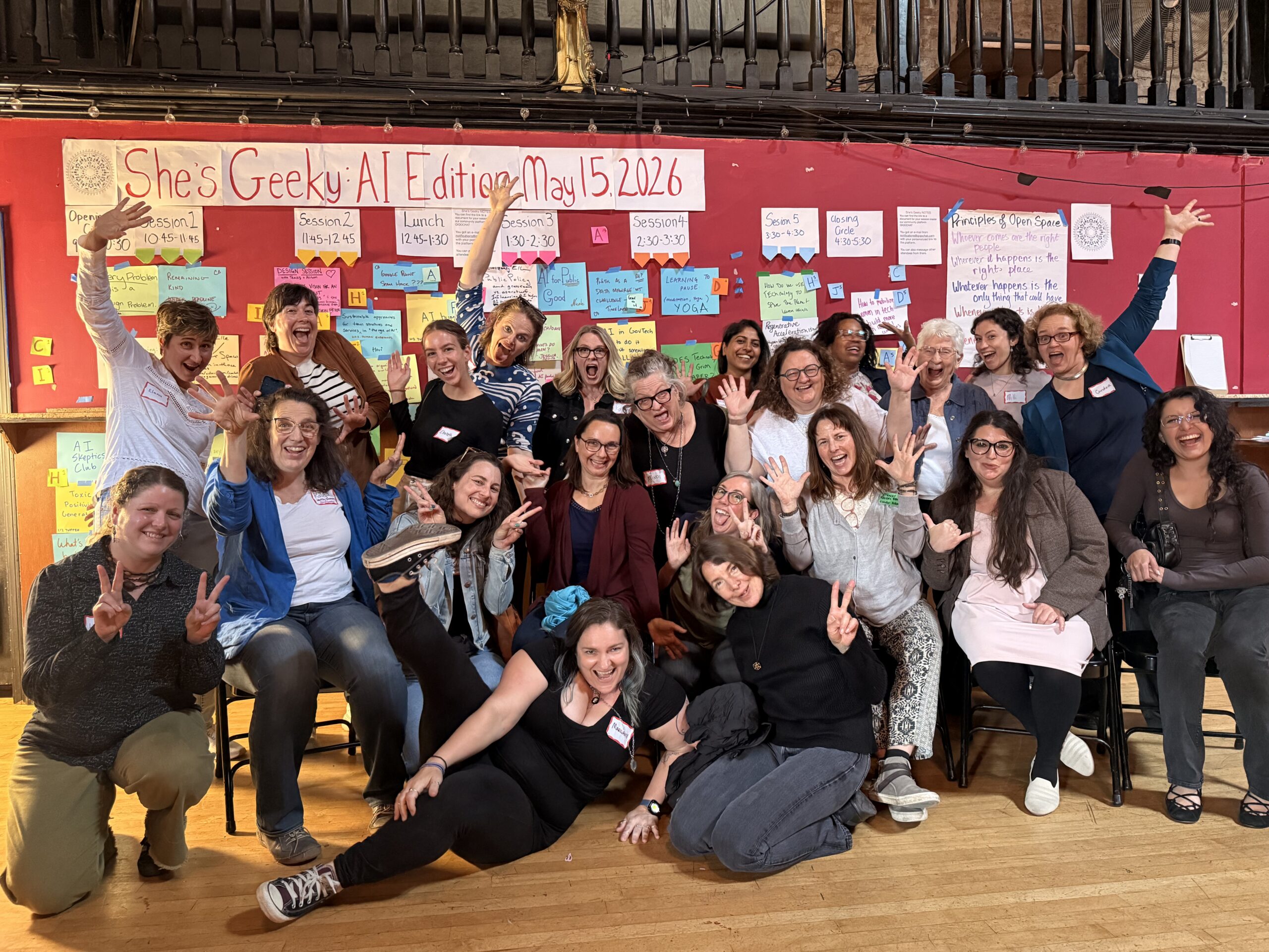 A large group of women at She's Geeky AI Edition, arms raised, in front of the Open Space session board.