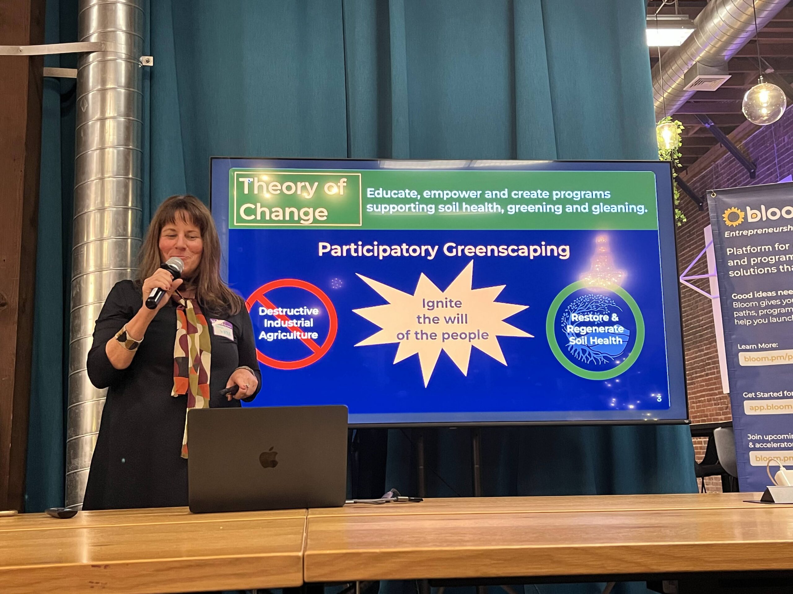 Tracey presenting on stage, microphone in hand, beside a slide reading Theory of Change: participatory greenscaping, ignite the will of the people, restore and regenerate soil health.