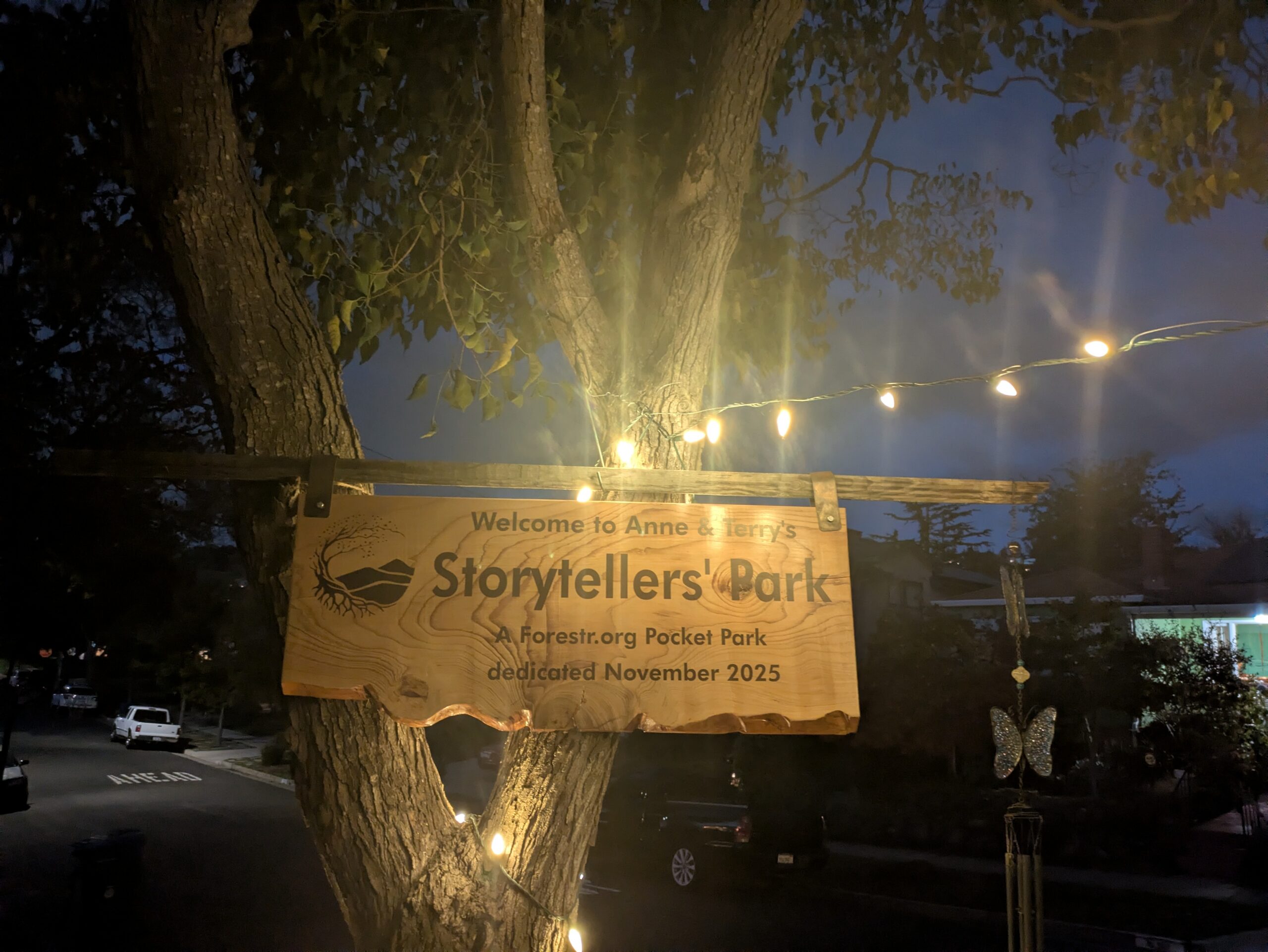 A hand-carved wooden sign reading Storytellers' Park hanging from a tree at dusk, lit by string lights.